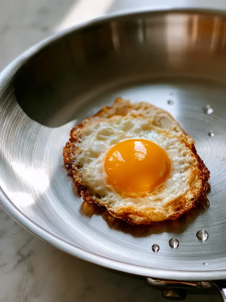 High-angle view of a fried egg with a glossy orange yolk in a stainless steel pot on a marble background.